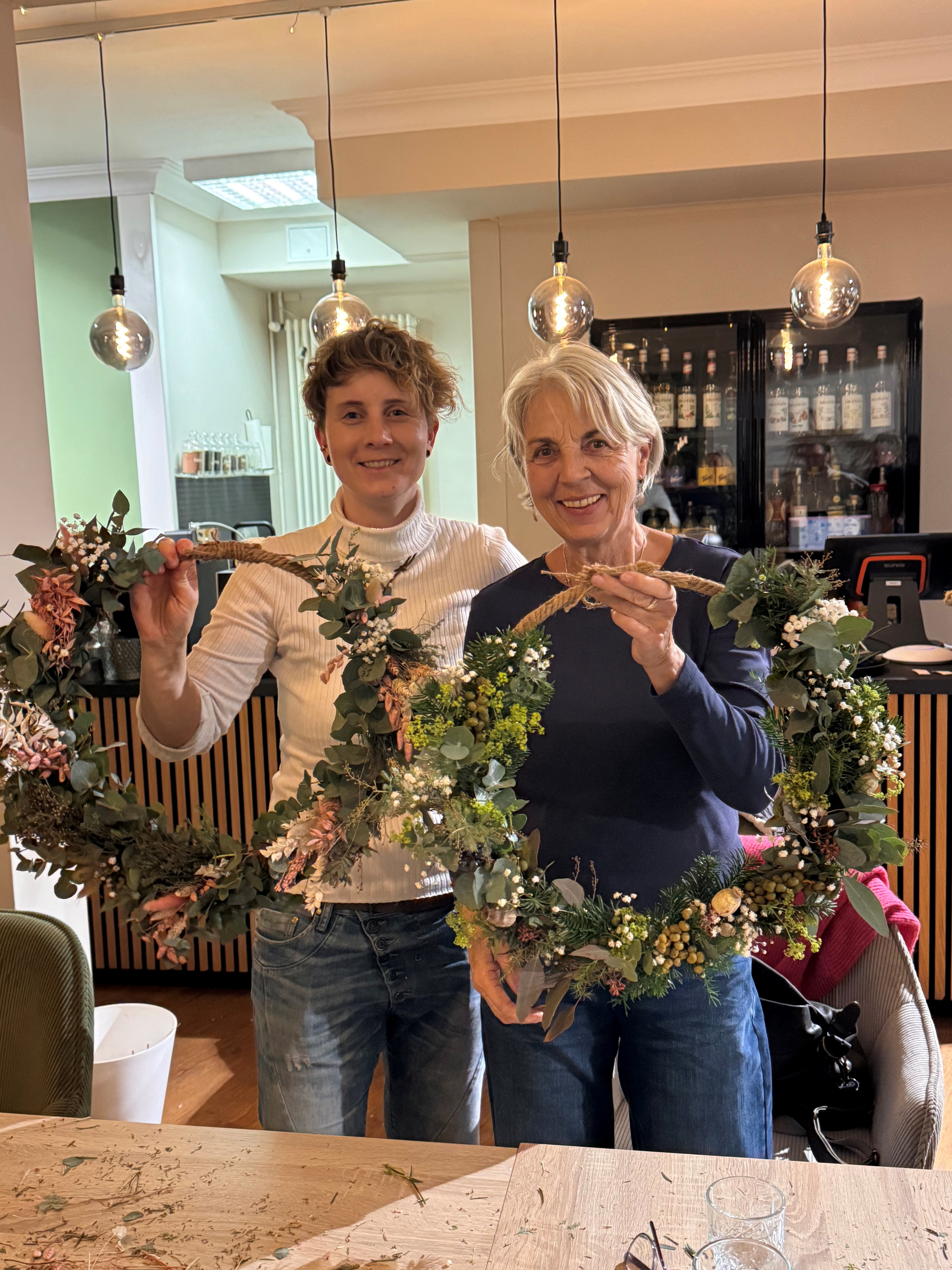 Zwei lächelnde Frauen präsentieren stolz ihre selbstgemachten, festlichen Winterkränze aus frischem Grün und Trockenblumen, entstanden beim kreativen Teambuilding 'Zauberhafte Vorweihnachtszeit' bei Ly's.