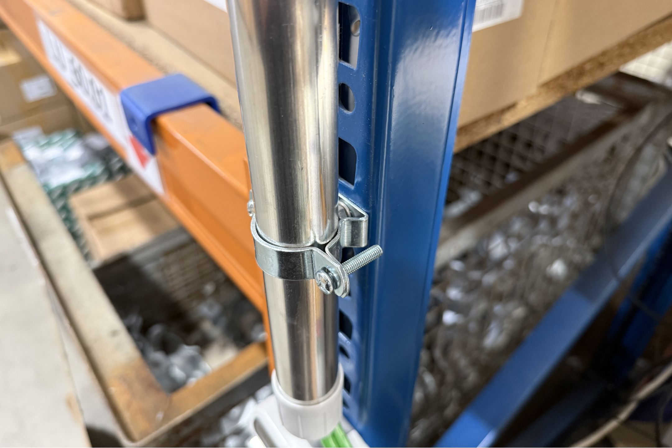 Kabelschellen-Metallklemme befestigt ein blankes Rohr sicher an einem blauen Lagerregal. Die verzinkte Schelle ist mit Schraube und Mutter montiert.
