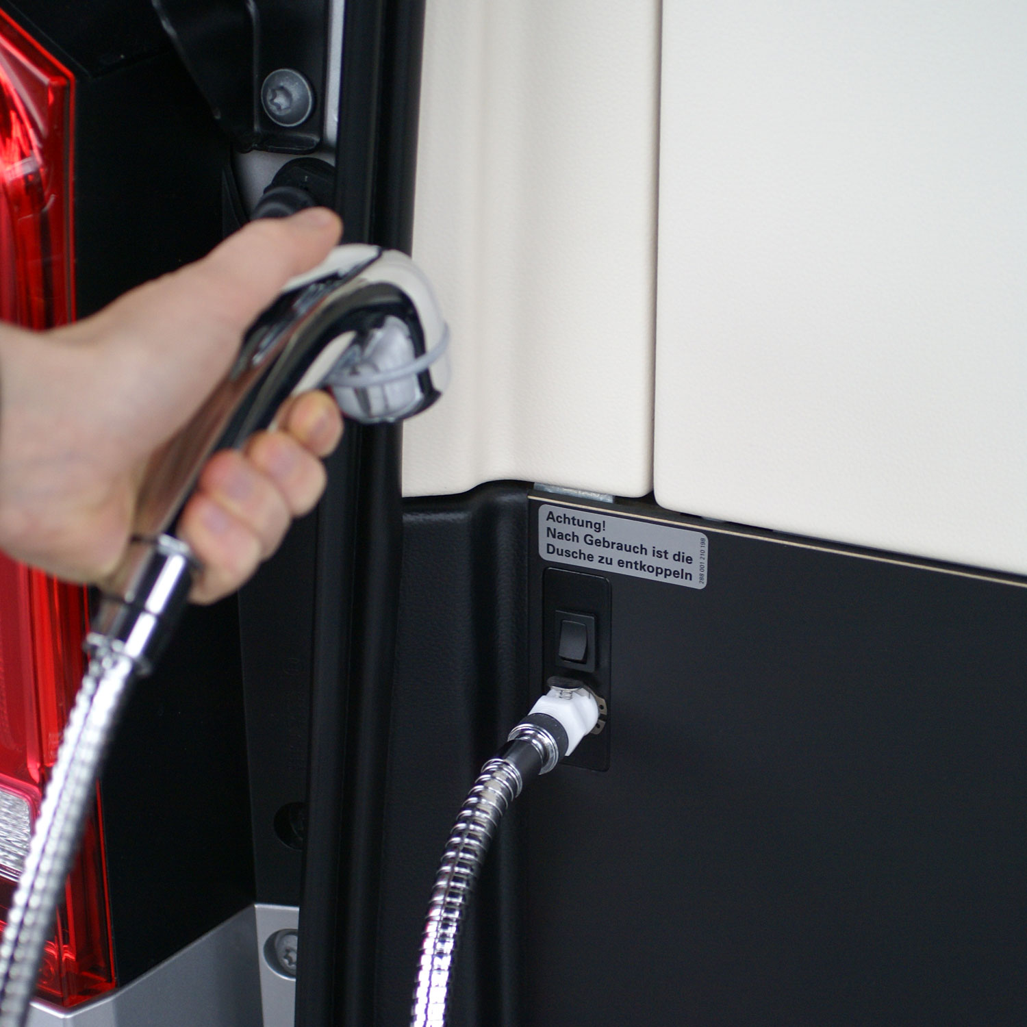 Hebeldusche Camping: Nahaufnahme des Duschkopfes, der von einer Hand gehalten und an den praktischen Entnahmeport im Fahrzeug angeschlossen wird. Eine Hand hält den glänzenden Duschkopf der Hebeldusche Camping, deren flexibler Metallschlauch an einen externen Wasseranschluss im Fahrzeugheck angeschlossen ist.