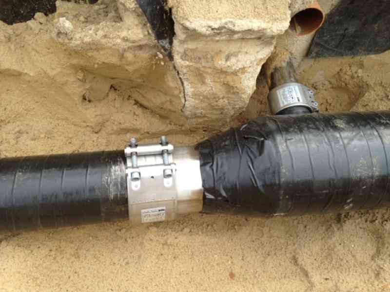 Rohrverbinder, Rohrkupplung, Rohre verbinden: Zwei schwarze Rohre werden im sandigen Boden durch eine robuste Metallkupplung mit Schrauben verbunden; ein Abschnitt der Verbindung ist mit schwarzem Isolierband umwickelt.