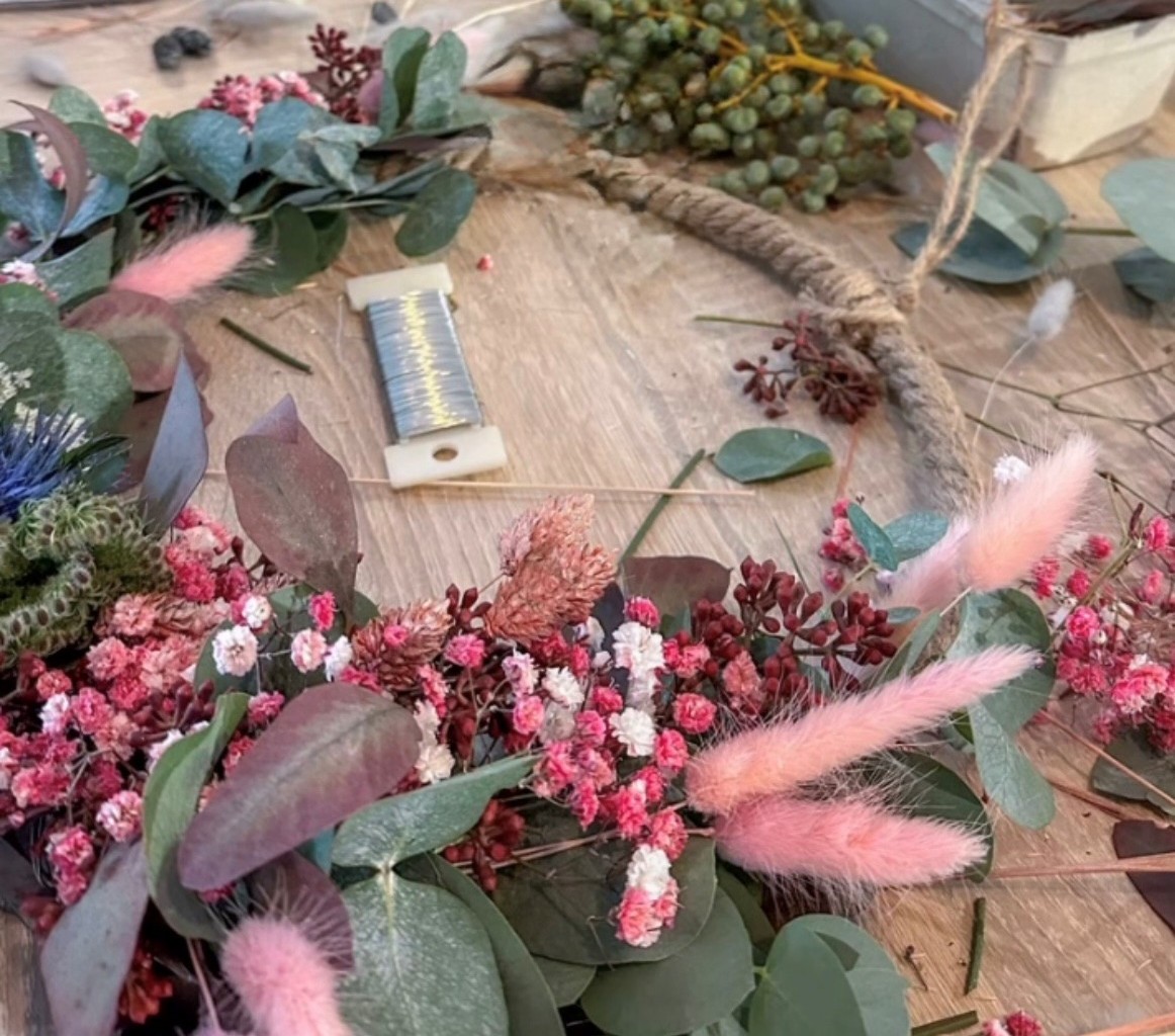 Nahaufnahme von Bastelmaterialien für einen festlichen Kranz auf einem Holztisch, mit Eukalyptusblättern, kleinen rosa und weißen Blüten, roten Beeren und flauschigen rosa Gräsern. Ein Spule Draht und Bindfaden sind ebenfalls sichtbar. Dieses Bild fängt d