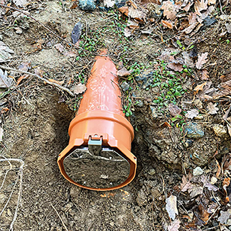 Orangefarbene Rückstauklappe / Froschklappe für Spitzenden / Rohrenden mit sichtbarer silberner Klappe, teilweise in erdigen Boden mit trockenen Blättern und grünen Pflanzen eingebettet.