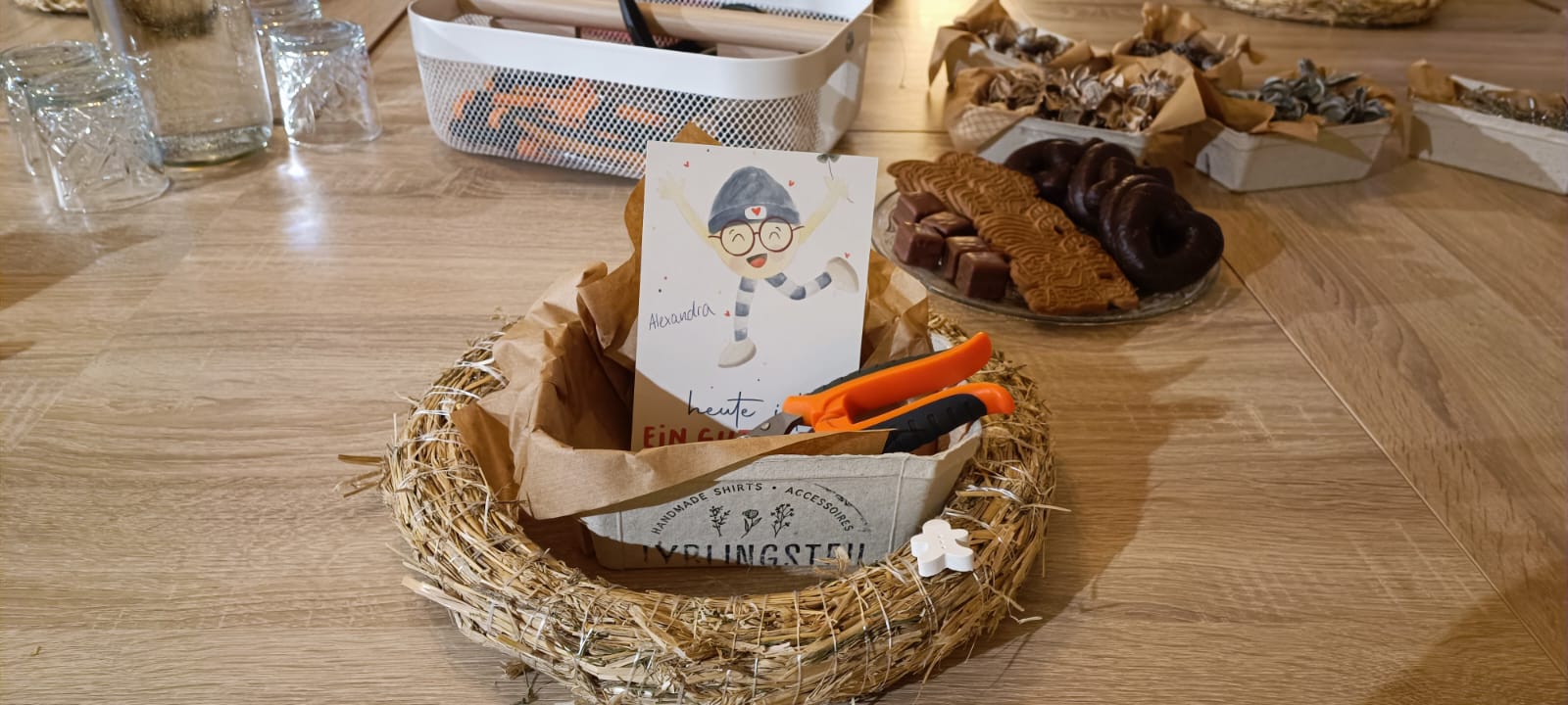 Detailaufnahme eines weihnachtlichen Bastelsets für das kreative Teambuilding bei Ly’s, mit Strohkranz, Bastelschere, personalisierter Karte für Alexandra und festlichem Gebäck.
