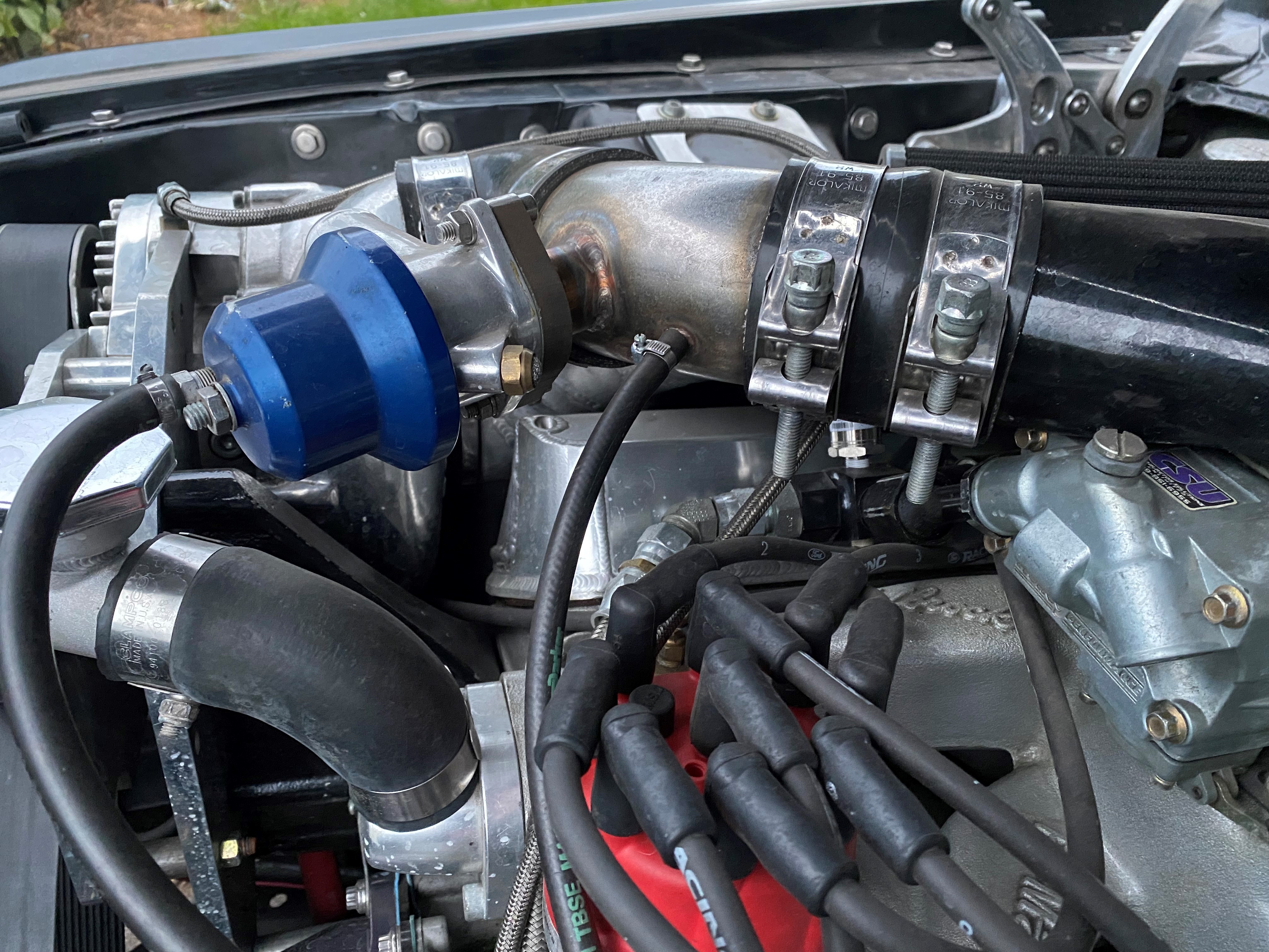 Detailansicht des Motorraums eines Fahrzeugs mit verschiedenen Motorkomponenten. Im Vordergrund sind schwarze Zündkabel, silberne Aluminiumteile, Schläuche und Rohrverbindungen zu sehen. Besonders auffällig sind zwei robuste Gelenkbolzenschellen (Mikalor